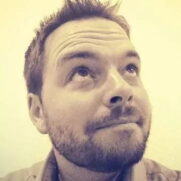 A man with short hair and a beard looks up and slightly to the side with a thoughtful expression. The image has a sepia tone.