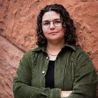 A person with curly hair and glasses, wearing a green jacket over a black shirt, stands with arms crossed against a textured, reddish-brown wall.